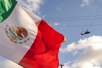 Primer plano de una bandera mexicana ondeando con un helicóptero oscuro y líneas eléctricas bajo un cielo azul parcialmente nublado