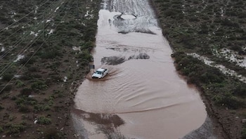 Una pala mecánica amarilla limpia barro en una carretera mojada junto a vehículos y una persona con impermeable