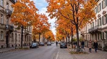 Una calle con edificios residenciales, automóviles estacionados, personas caminando y árboles de hojas anaranjadas.