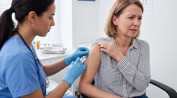 Una profesional de la salud con uniforme azul y guantes azules inyecta el brazo de una persona adulta con camisa de rayas, que toca un apósito.