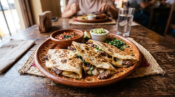 Tres quesadillas de carne y queso en un plato de barro con salsa roja, guacamole, cilantro y un vaso de agua sobre una mesa de madera.