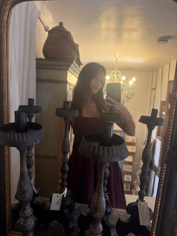 Reflejo de una mujer en un espejo, vestida de oscuro, tomando una selfie con un teléfono móvil. En primer plano, tres candelabros de metal antiguos