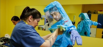 Una persona en scrubs azules asiste a otra poniéndose un traje de protección biológica azul con guantes morados y visor transparente en una sala amarilla