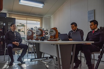 Tres hombres sentados en una mesa con tres medidores de energía eléctrica en soportes de madera; dos hombres usan laptops, el tercero escucha