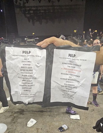 Una persona sostiene un setlist arrugado del concierto de Pulp con el listado de canciones para el Palacio de los Deportes. Al fondo, se ve un escenario con equipo y técnicos