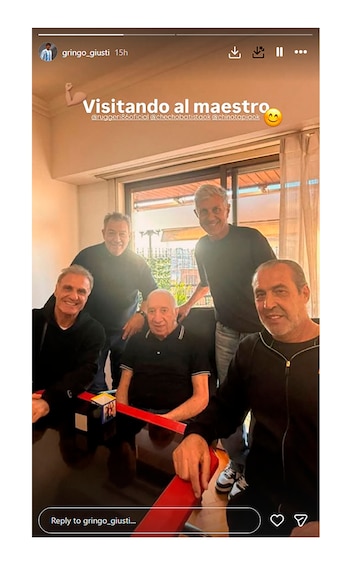 Cinco hombres en una habitación iluminada, uno sentado al centro y cuatro de pie alrededor de una mesa