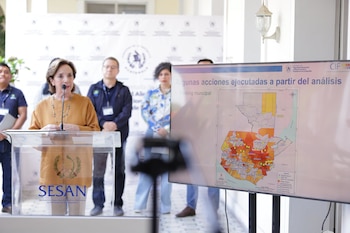 Mujer en un podio transparente habla a un micrófono, junto a otras personas, frente a una pantalla con un mapa de Guatemala y texto
