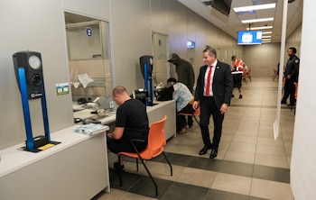 Dariel Fernandez, un hombre en traje oscuro y corbata roja, camina sonriente por una oficina moderna con ventanillas de atención al público y clientes