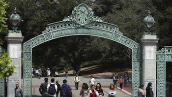 Na frente está o Sather Gate, um arco verde brilhante da Universidade da Califórnia, com pessoas andando por baixo e ao redor dele.