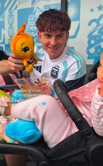 Hombre sonriente con camiseta de la selección argentina sostiene un muñeco de abeja de peluche junto a un bebé en cochecito con fondo azul y blanco