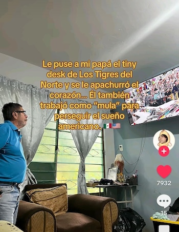 Un hombre de pie con camisa azul mira un televisor encendido en una sala con sillón, cortinas y texto superpuesto en pantalla