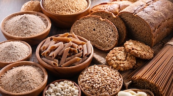 Pan integral rebanado, fideos integrales, galletas de avena, harina integral en cuencos, granos de trigo, y legumbres blancas en recipientes de madera sobre una superficie oscura.