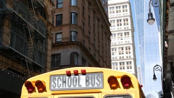Vista trasera de un autobús escolar amarillo con edificios altos de ladrillo y un rascacielos de cristal al fondo. Se aprecian escaleras de incendio y una farola
