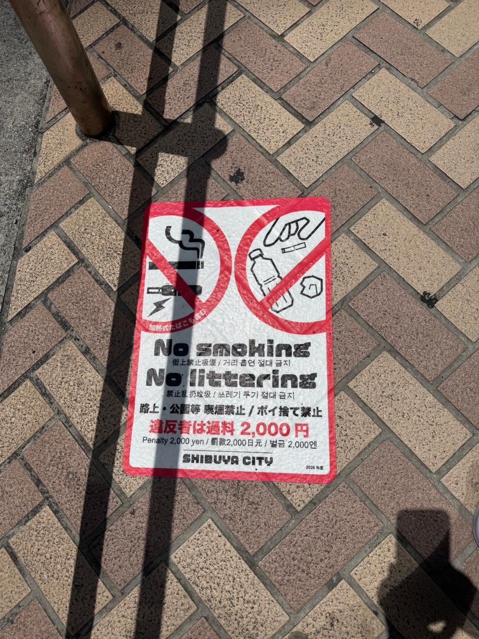 Un cartel en una acera de Shibuya, Japón, advierte a los transeúntes sobre multas de 2.000 yenes por fumar y tirar basura en la vía pública, buscando mantener la limpieza urbana. (Infobae)