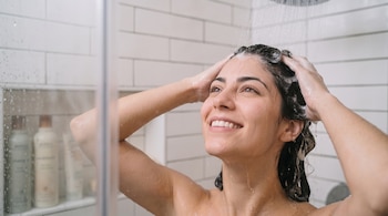 Una mujer lava su cabello con espuma bajo el agua de la ducha en un baño con azulejos blancos.