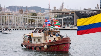 Barco decorado con banderas y personas en el puerto de Barcelona con edificios al fondo y un puente. Una bandera colombiana grande se superpone a la derecha.