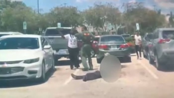 Una persona yace en el suelo de un estacionamiento junto a varios automóviles y agentes policiales