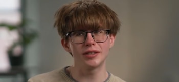 Primer plano de Matthew Lane, un joven con cabello castaño y gafas de montura negra, vistiendo un suéter beige. Su boca está ligeramente abierta como si estuviera hablando