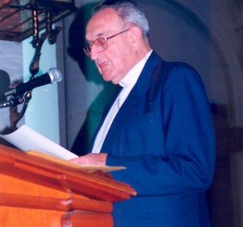 Retrato de Monseñor Juan José Gerardi, un hombre mayor con gafas y traje azul, hablando frente a un micrófono en un podio de madera con papeles
