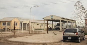 Exterior del Establecimiento de Ejecución Penal 5 Cipolletti con un edificio beige, rejas blancas, un cartel con el nombre y un auto plateado estacionado