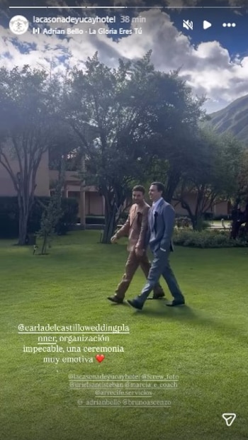 Dos hombres en traje, uno en marrón y otro en azul, caminan sobre césped verde con árboles, edificios y montañas al fondo bajo un cielo nublado