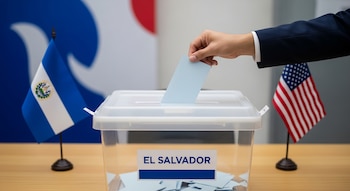 Mano deposita boleta en urna transparente con etiqueta 'El Salvador' sobre mesa de madera clara. Banderas de El Salvador y Estados Unidos desenfocadas al fondo.
