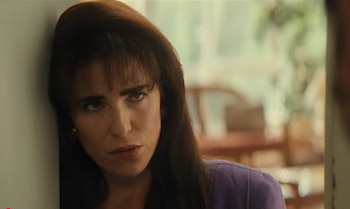 Mujer con cabello castaño oscuro y flequillo, vestida con una blusa morada, apoyada en una pared clara y mirando directamente a la cámara