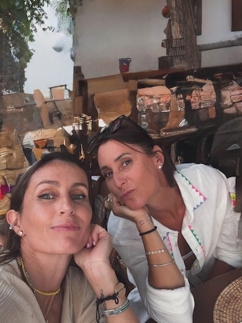 Dos mujeres adultas posan para la cámara, una con cabello largo y la otra con gafas de sol sobre su cabeza, ambas con joyas y vestimenta casual