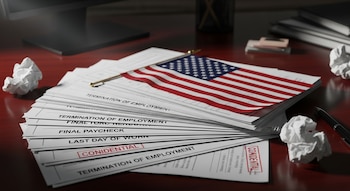 Primer plano de una pila de documentos de terminación de empleo sobre un escritorio de madera, con una pequeña bandera de Estados Unidos encima. También se ven papeles arrugados y un bolígrafo.