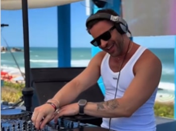 Un hombre con camiseta blanca de tirantes, gafas de sol y auriculares manipula una mesa de mezclas con el mar de fondo