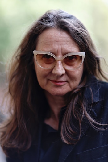 Retrato de Lucrecia Martel, mujer de mediana edad con gafas de montura clara y cabello castaño con canas. Viste una chaqueta oscura, mirando fijamente a cámara con un fondo verde borroso