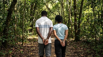 Hombre y mujer de espaldas en un bosque denso; ambos tienen las manos atadas con cuerda detrás. Caminan por un sendero con luz solar filtrada.