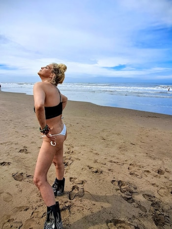 Leticia Brédice de perfil, con un bikini negro y blanco, botas negras con tachas, posando en una playa de arena frente al mar bajo nubes