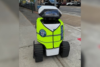 Un robot blanco con un chaleco reflectante amarillo se encuentra sobre una acera de cemento, con vehículos estacionados al fondo