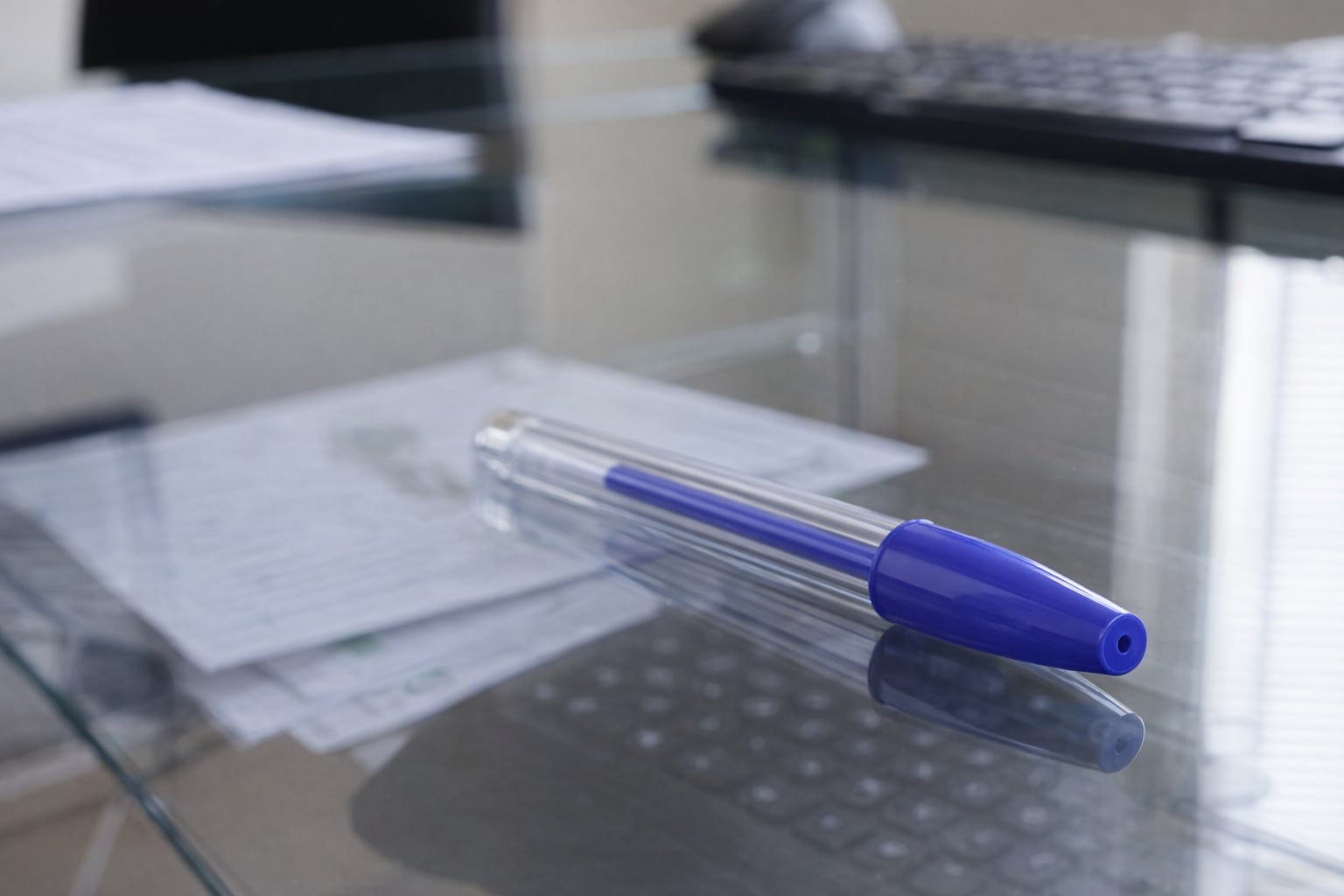 Un lapicero azul con su tapa puesta y el orificio de seguridad visible descansa sobre una mesa de cristal, reflejando documentos y un teclado en un ambiente de oficina. (Imagen Ilustrativa Infobae)