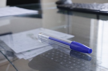 Primer plano de un lapicero azul con tapa transparente y orificio de ventilación, colocado sobre una mesa de cristal con reflejos de papeles y un teclado.