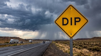 Una carretera con varios vehículos, una señal de tránsito amarilla con la palabra 'DIP' en primer plano, y un cielo cubierto de nubes de tormenta con lluvia.