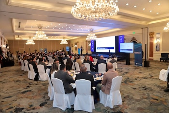 Amplio salón con techo alto, candelabros, múltiples mesas redondas con sillas blancas ocupadas por asistentes, tres pantallas grandes en el escenario