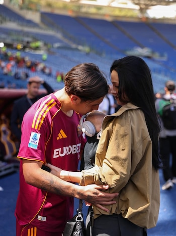 Paulo Dybala, com camisa da Roma, se inclina para abraçar Oriana Sabatini, que segura um bebê com fones de ouvido no campo de futebol.