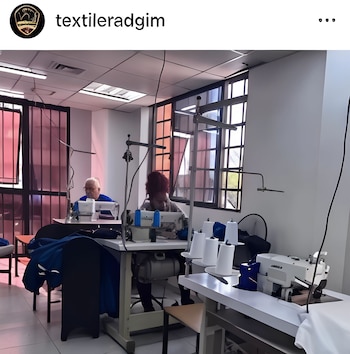 Dos personas cosiendo en máquinas industriales dentro de un taller luminoso. Un hombre mayor a la izquierda y una mujer a la derecha, rodeados de carretes de hilo
