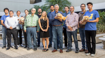 Once personas diversas posan en un patio exterior soleado, algunos sosteniendo peluches de perro. Hay un edificio claro y palmeras al fondo