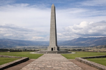 Un obelisco de concreto gris en el centro de un área verde con caminos de piedra, montañas al fondo y un cielo con nubes dispersas.