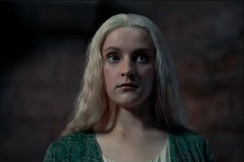 Mujer joven de cabello rubio platinado y ojos claros, vestida con un atuendo verde con patrones, mira hacia arriba sobre un fondo oscuro