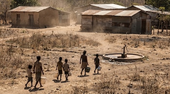 Un grupo de personas, incluidos niños, camina por un paisaje árido con baldes vacíos hacia un pozo seco. Se ven casas de adobe y lámina bajo una luz solar intensa.