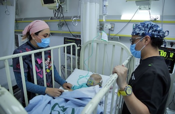 Una enfermera y un médico con mascarillas quirúrgicas examinan a un niño con máscara de oxígeno acostado en una cuna hospitalaria, rodeados de equipos médicos