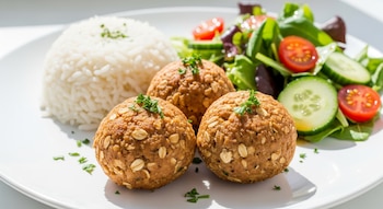 Plato blanco con tres albóndigas doradas de atún y avena, arroz blanco cocido y una ensalada de lechuga, rodajas de pepino y mitades de tomate cherry.