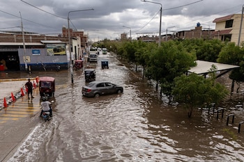 Una calle inundada con un automóvil gris, mototaxis, una motocicleta, árboles y edificaciones bajo un cielo cubierto de nubes