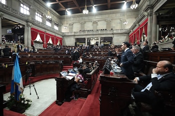 Vista panorámica de la sala de sesiones del Congreso de Guatemala. Legisladores sentados en pupitres de madera oscura, una bandera guatemalteca y pantallas