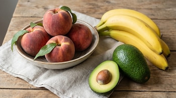 Melocotones en un cuenco sobre un paño de lino, un racimo de plátanos y dos aguacates (uno cortado a la mitad con el hueso visible) sobre una mesa de madera.