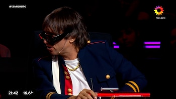 Hombre con gafas de sol oscuras y chaqueta militar azul marino con detalles dorados y rojos, camiseta blanca, sentado en un set oscuro con luces rojas